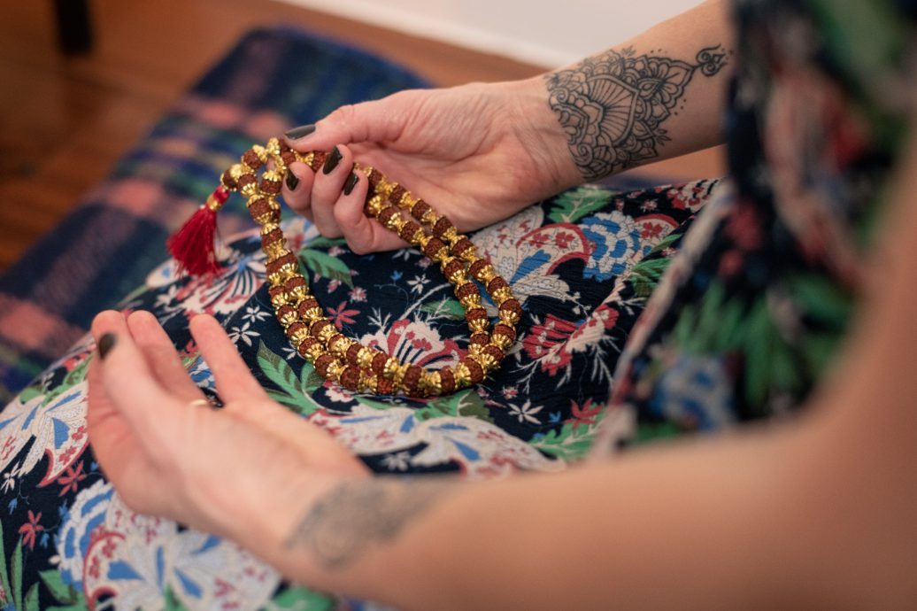 Person with tattooed arms holding a golden beaded mala necklace over a colorful floral fabric during a meditation or yoga practice session.
