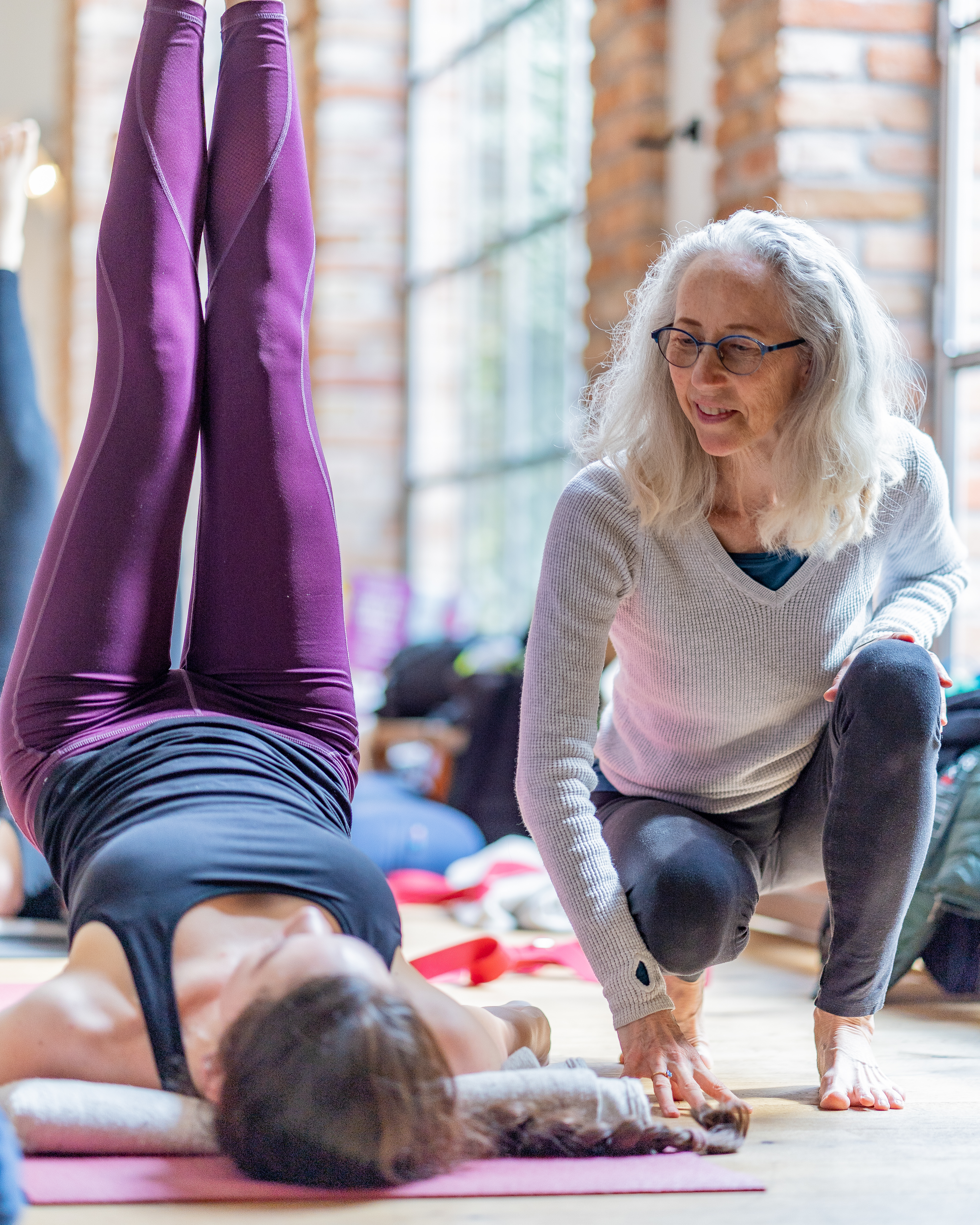 Yoga instructor with white hair and glasses observing a student performing a headstand on a mat in a bright studio with brick walls.