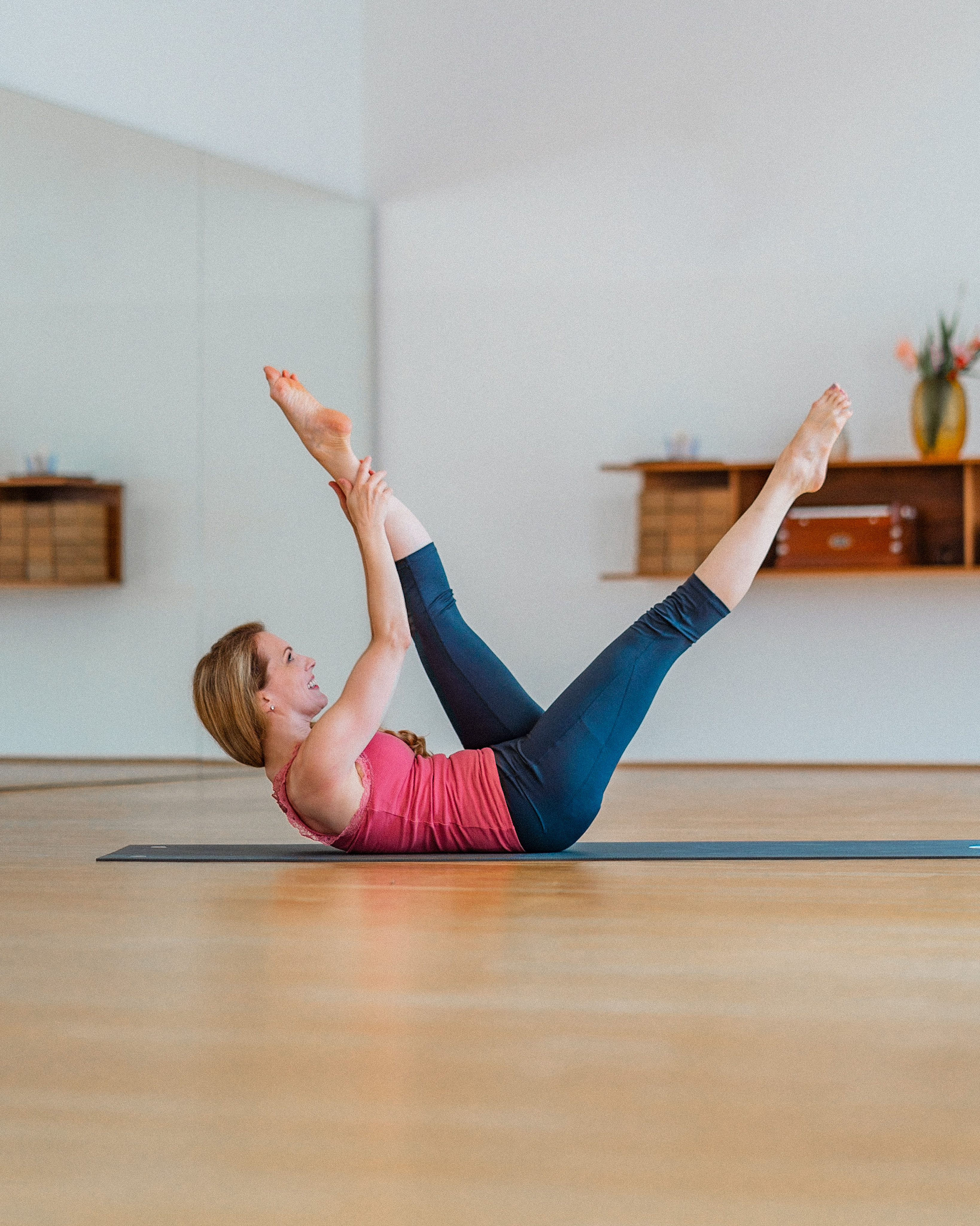 Person performing a pilates or yoga exercise on a mat in a bright studio with wooden shelves and plants in the background.