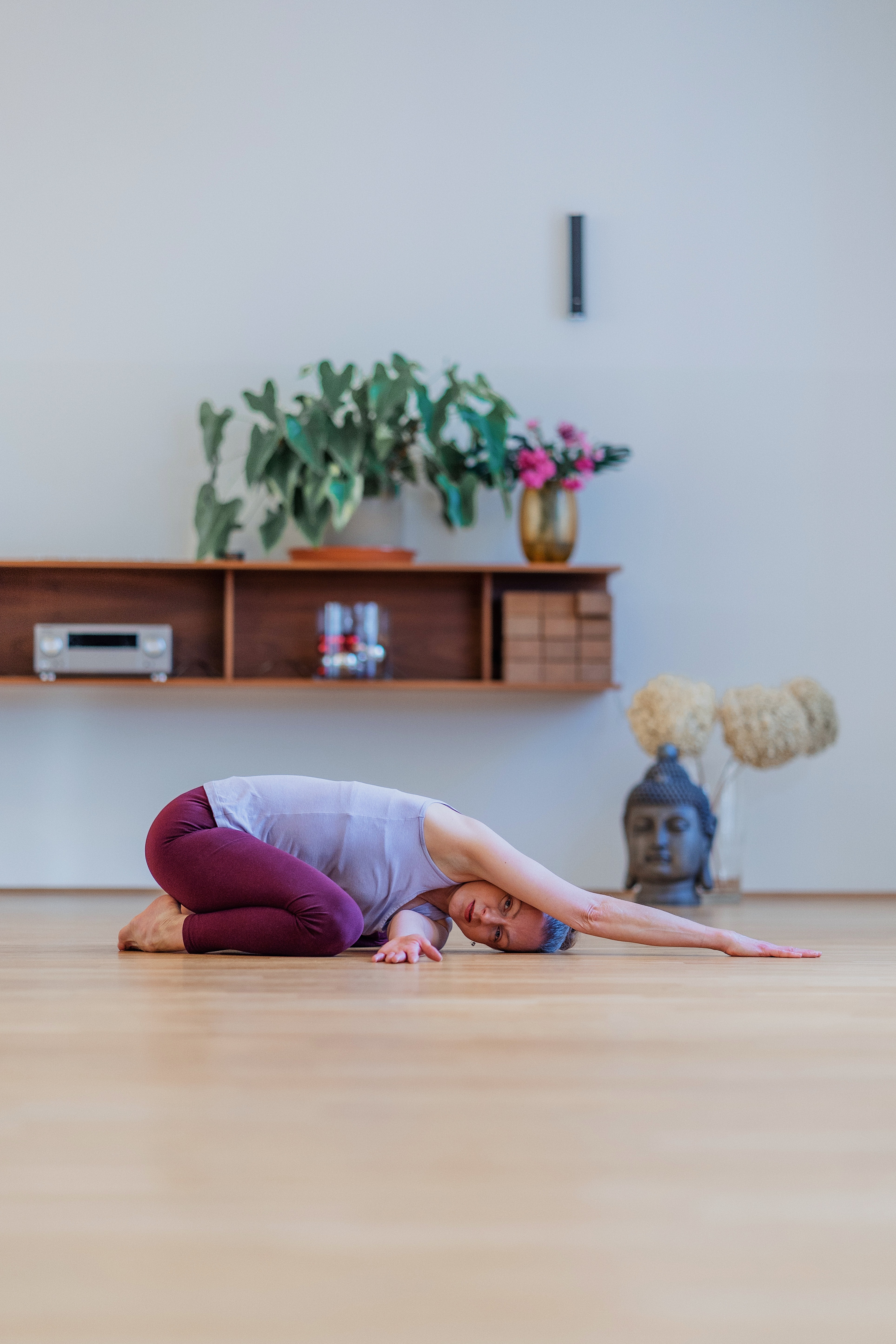 Person performing a forward fold yoga pose on a wooden floor in a bright studio with plants, Buddha statue, and wall-mounted shelving in the background.