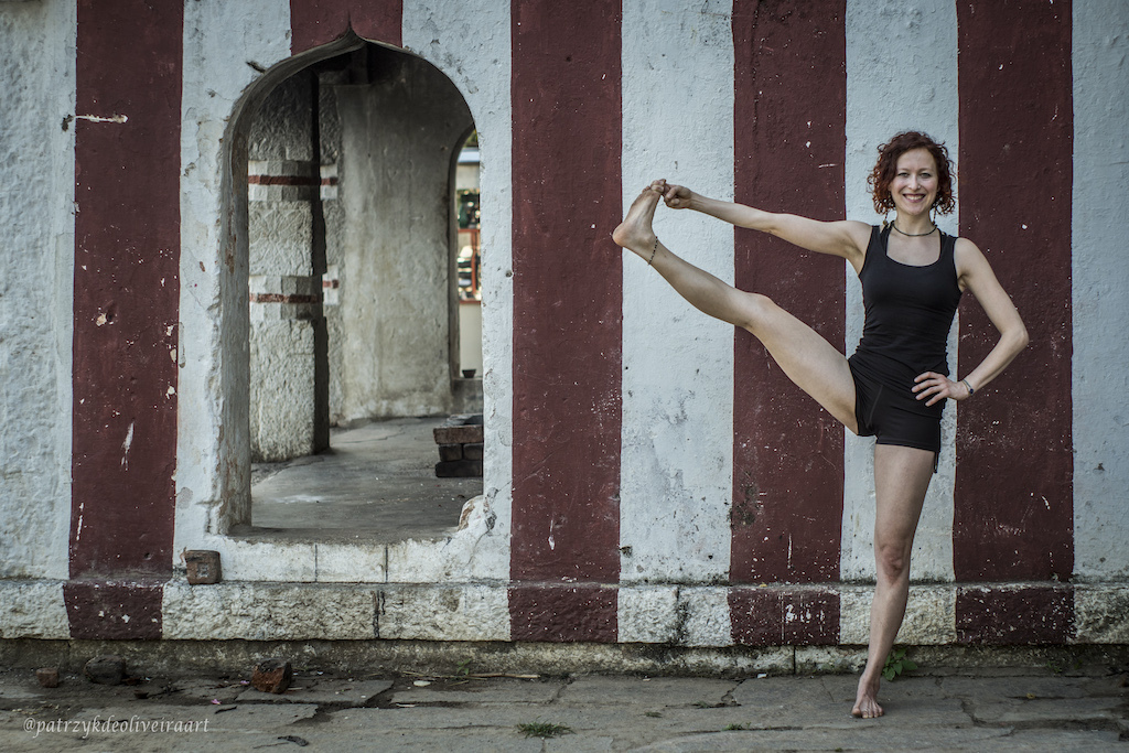 Dancer in black outfit performing a standing leg extension pose against striped architectural columns with an arched doorway in background.