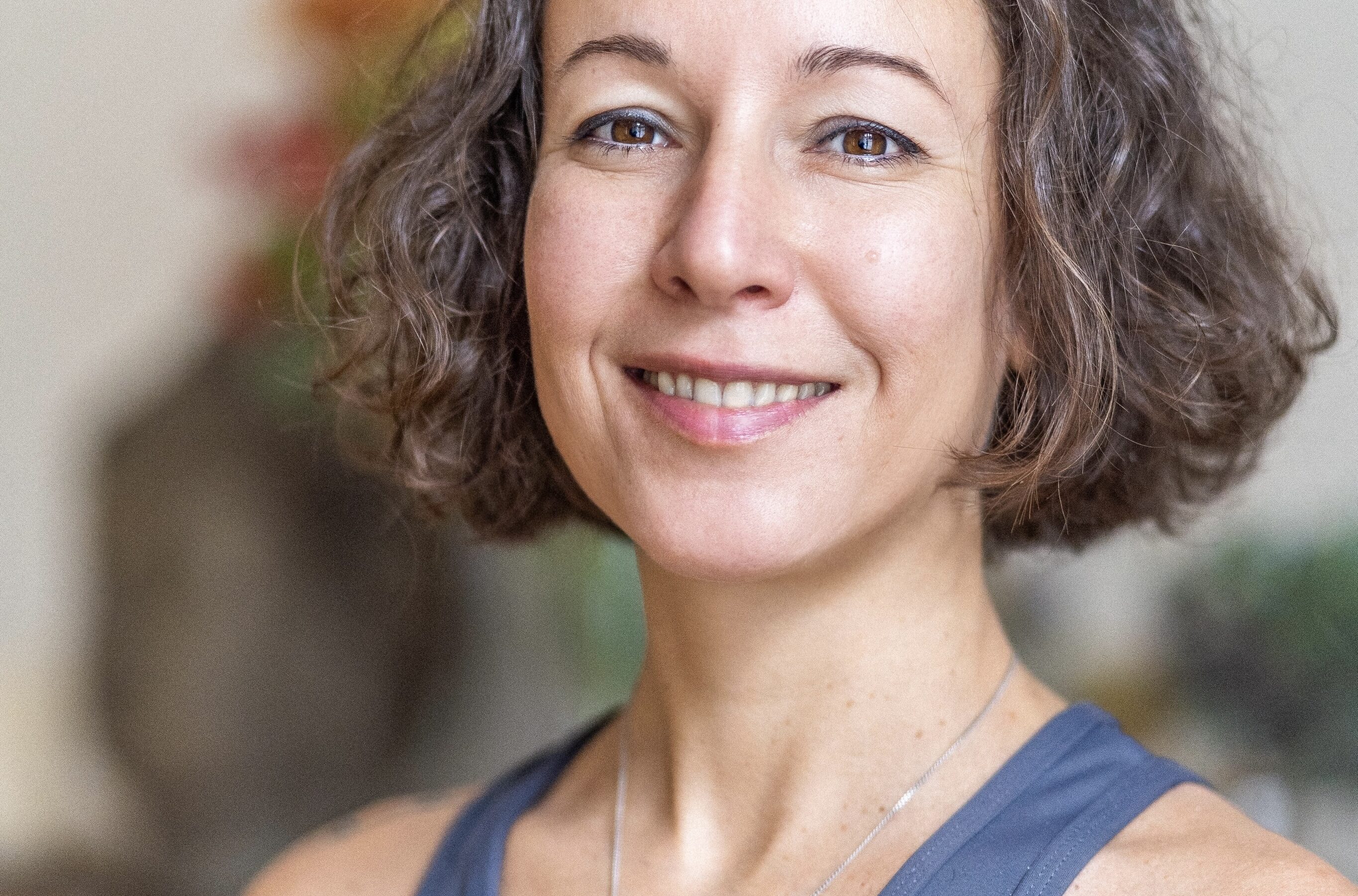 Person mit schulterlangem braunem Haar, lächelnd, trägt blaues Yoga-Top mit silbernem Anhänger, vor unscharfem Hintergrund