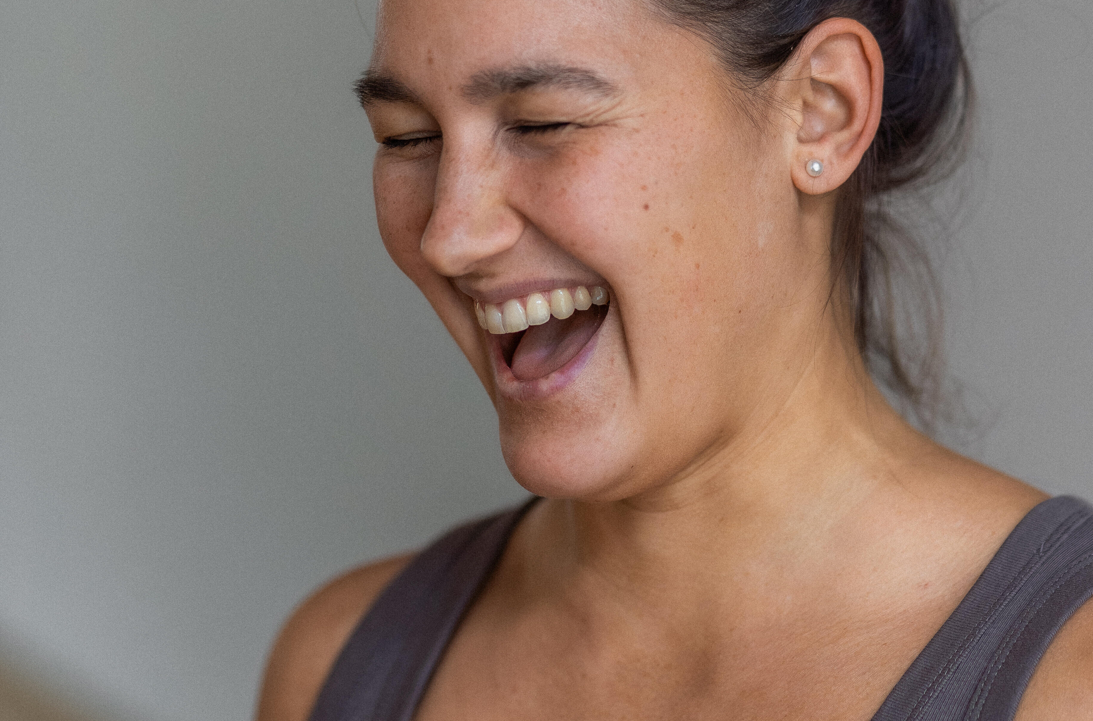 Person with dark hair in a top bun, wearing a gray tank top, smiling with eyes closed against a neutral background