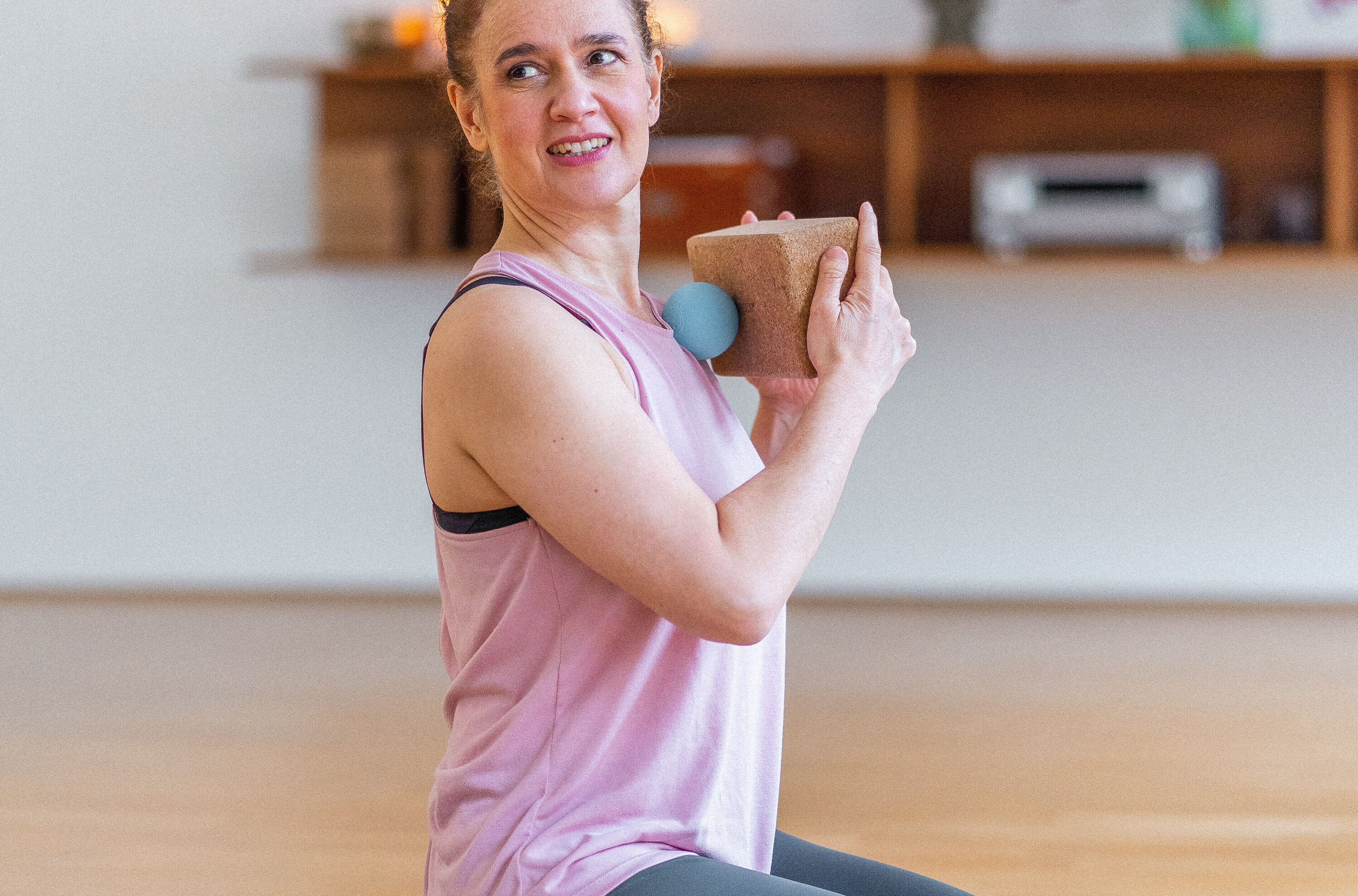 Person in pink tank top and gray leggings performing a yoga pose while holding a cork block during a fitness class in a bright studio.